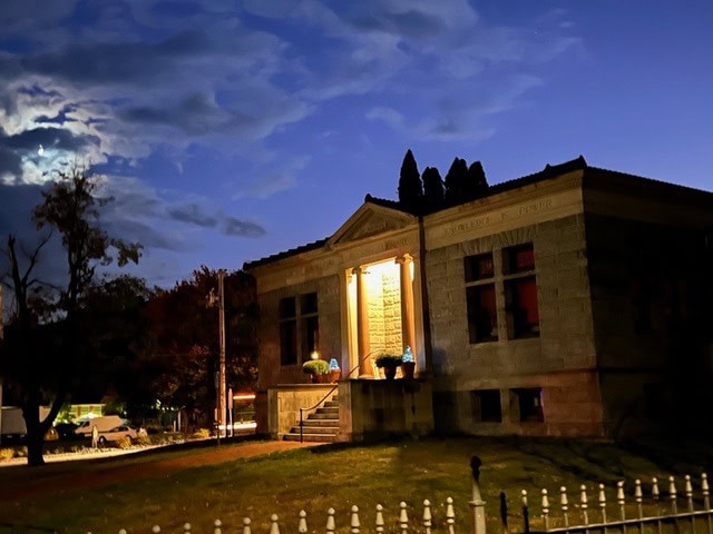 stone building with columns and illuminated entrance at dusk, surrounded by trees and a grassy lawn, with a cloudy sky and moon visible in the background.