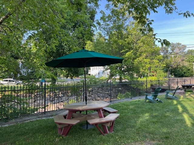 a wooden picnic table with attached benches and a green umbrella sits on grass near a black metal fence, with trees, adirondack chairs, and houses in the background.