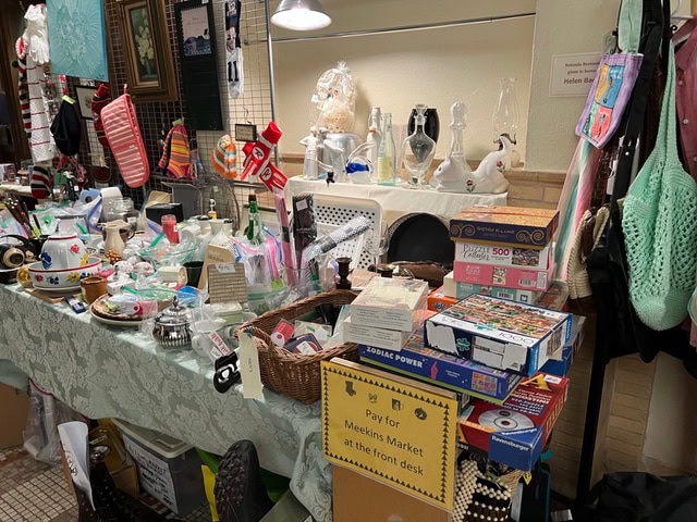 table at a market displaying assorted items including board games, glassware, baskets, teapots, and holiday stockings, with a sign instructing shoppers to pay at the front desk.