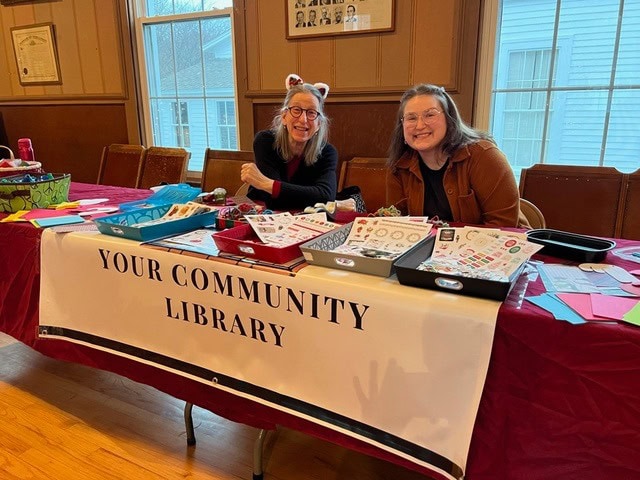 two people sit behind a table with arts and crafts supplies at a library event, with a banner reading "your community library" displayed in front of them.