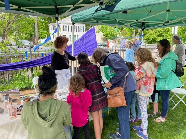 a group of people, including children and adults, gather under a canopy at an outdoor event, engaging with a woman at a display table.