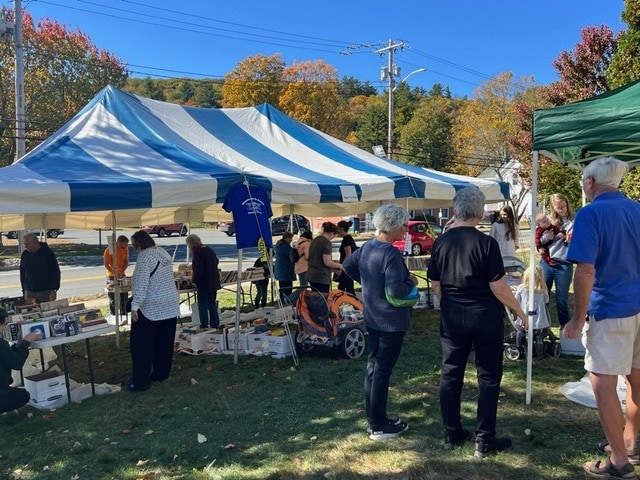 people gather under a blue and white striped tent at an outdoor event, looking at tables with items for sale on a sunny day with autumn trees in the background.