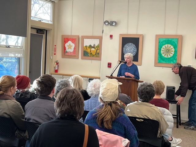 a woman speaks at a podium to a seated audience in a room decorated with colorful artwork; a man adjusts equipment nearby.