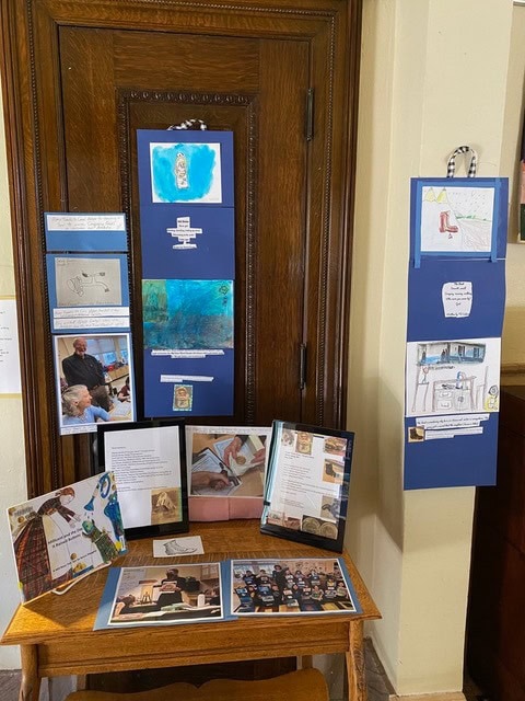 a display of student artwork, photographs, and written projects arranged on blue boards and a wooden table against a wooden door.