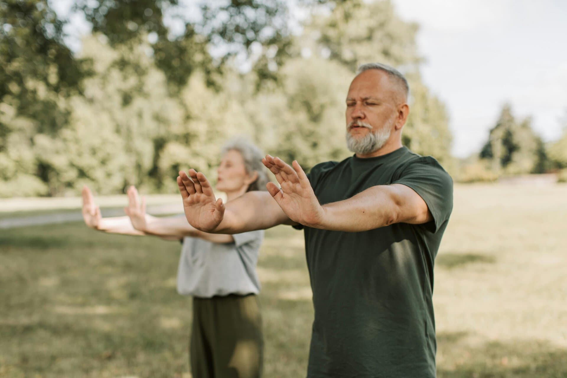 two older adults stand outdoors in a park, practicing a slow movement exercise with their arms extended forward, palms facing outward.