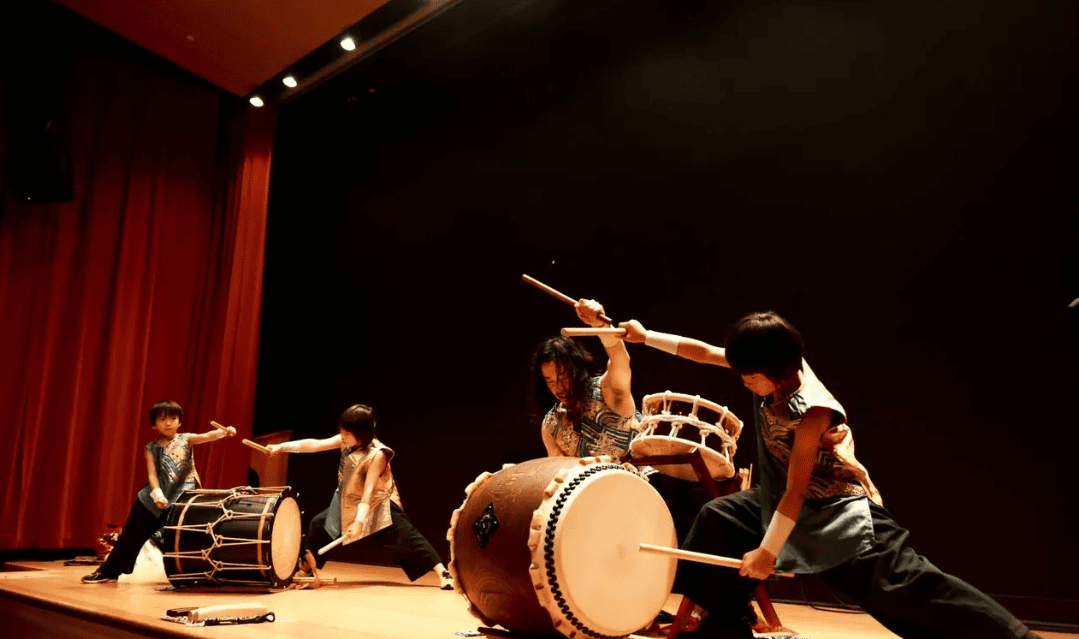 four people in matching outfits play traditional japanese taiko drums on a stage with dramatic poses and lighting.