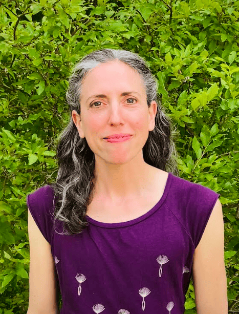 a woman with long, wavy gray and brown hair wearing a purple patterned top stands in front of green leafy bushes, looking at the camera with a neutral expression.