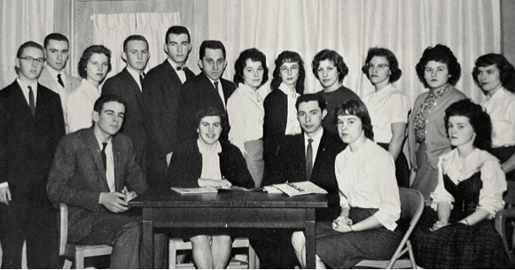 a group of sixteen young adults, dressed in formal attire, poses indoors around a table with papers, in front of a curtain.