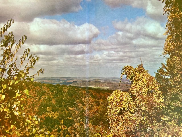 rolling hills covered with green and yellow foliage under a partly cloudy sky, with distant landscapes visible on the horizon.