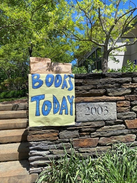 a large hand painted sign reading "books today" is taped to a stone wall next to steps leading up to a building at address 2003; paper bags are set on top of the wall.