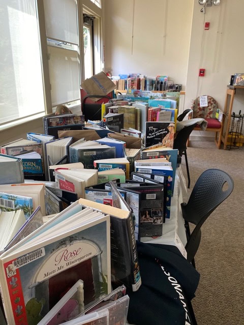 numerous books are displayed standing upright and fanned open on tables in a well lit room, with chairs around and more books stacked in the background.