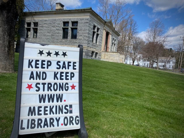 a sign in front of a stone building reads, "keep safe and keep strong www.meekinslibrary.org," standing on a grassy lawn under a partly cloudy sky.
