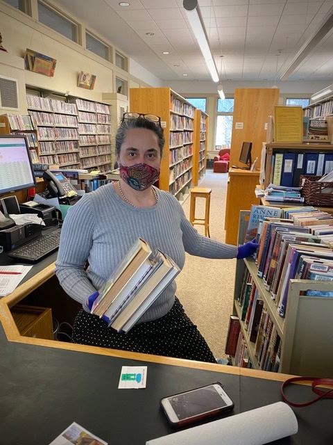 a person wearing a mask and gloves holds books while standing behind a library desk, with bookshelves and a computer visible in the background.