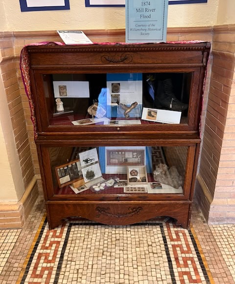 a wooden display case with glass doors exhibits historical artifacts and photographs, arranged on two shelves, against a tiled wall in a museum or historical society.