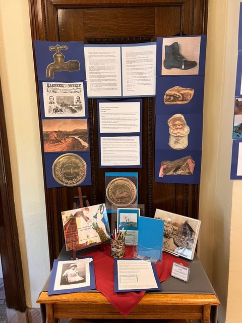 a display board with historical images, documents, and artifacts on a table, including books, pencils, postcards, and printed photos arranged neatly.