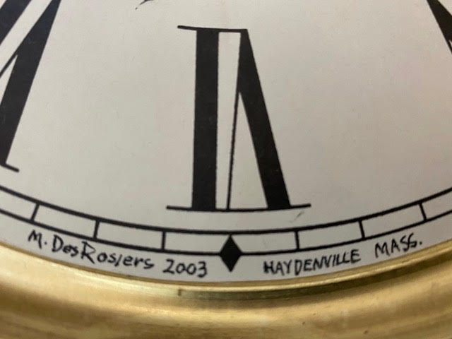 close up of a clock face showing part of the number six, with the inscription "m. desrosiers 2003 haydenville mass." written along the bottom edge.