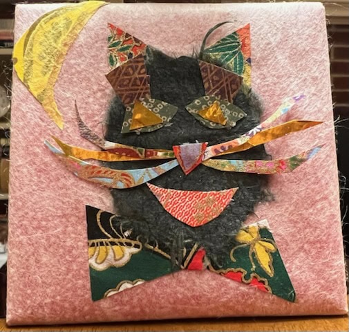 a collage artwork of a cat made from various patterned and textured fabrics on a pink background. the cat has large yellow eyes, multicolored whiskers, and a red tongue.
