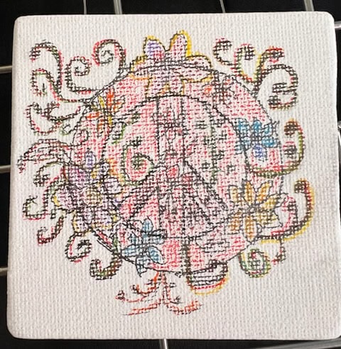a cross stitched pattern on white canvas features a central peace sign surrounded by colorful swirls and flowers.