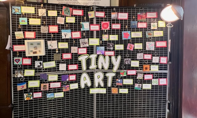 a display board labeled "tiny art" showcases many small square artworks and keychains, each with colored labels, under two spotlights.