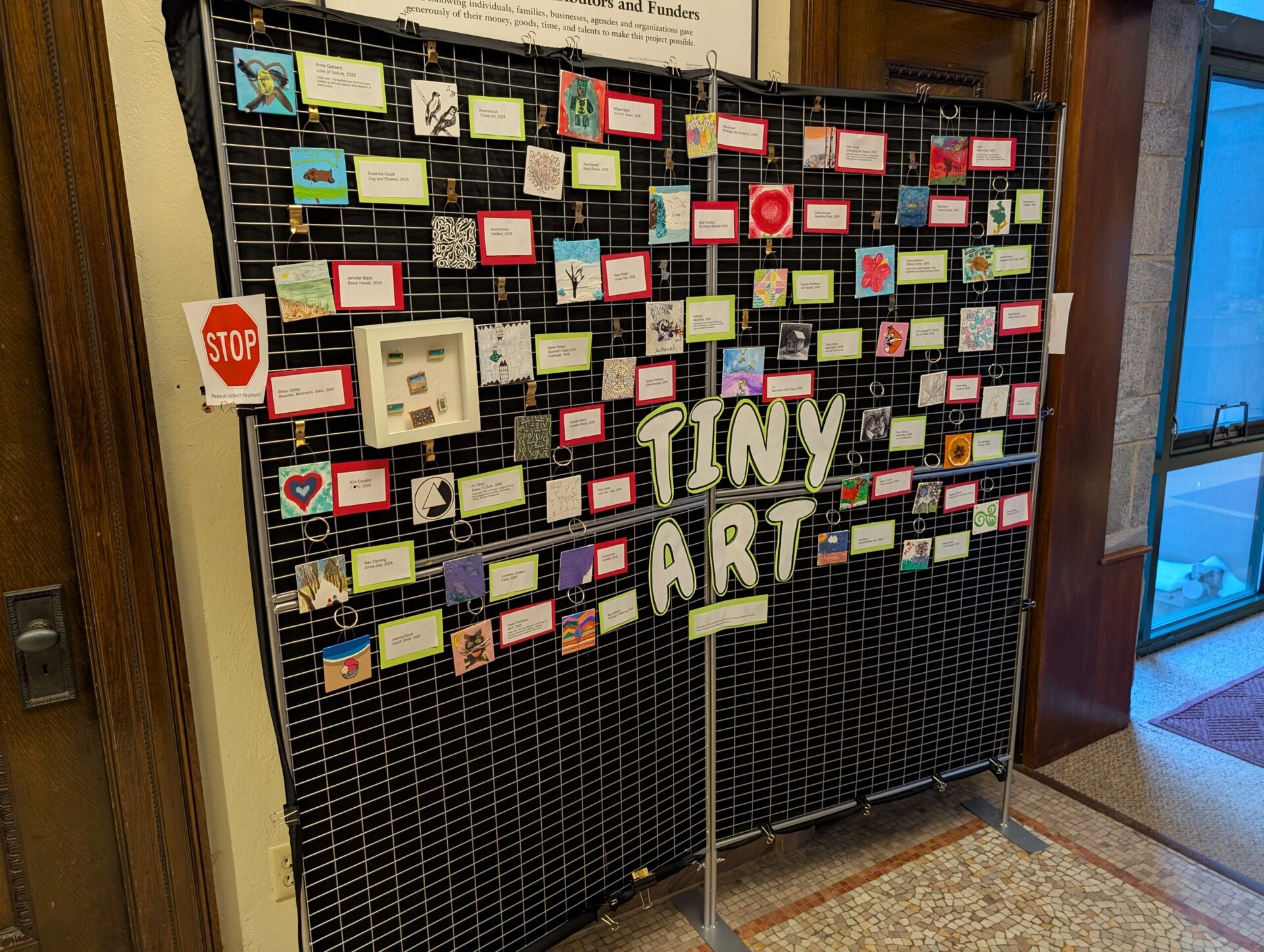 a display titled "tiny art" features numerous small artworks and colorful labels mounted on a black wire grid inside a building near a door.