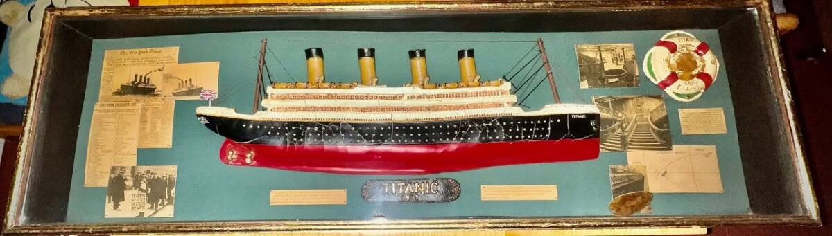 a framed display featuring a model of the rms titanic, surrounded by historical photographs, newspaper clippings, a life preserver replica, and informational plaques.
