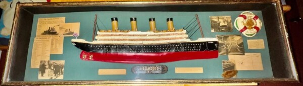 a framed display featuring a model of the rms titanic, surrounded by historical photographs, newspaper clippings, a life preserver replica, and informational plaques.