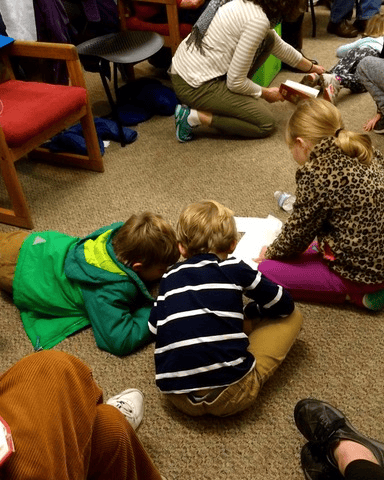 three children read or look at papers on the carpeted floor in a room, while two adults and another child are nearby.