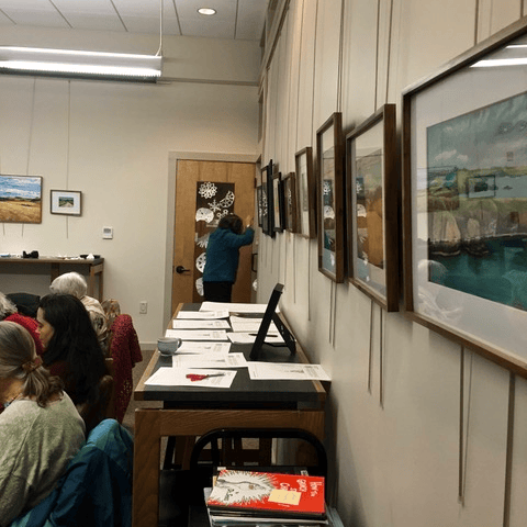 people sit and work at tables in a room with framed landscape paintings on the wall; one person stands near a door at the back, drawing or writing on it.