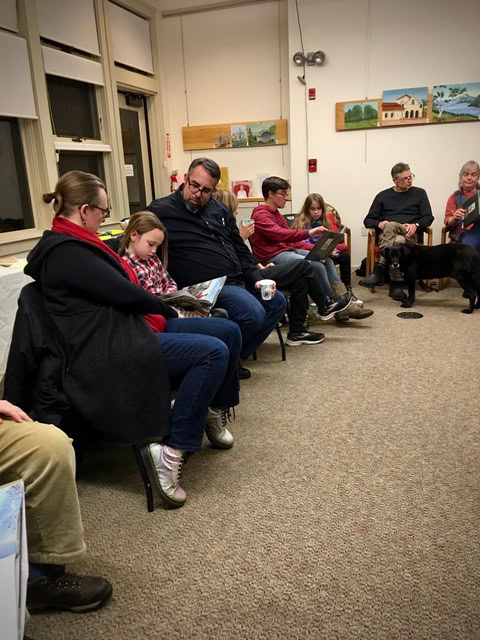 a group of people, including children and adults, sit in a circle in a room. some are reading books, and a dog is present. paintings hang on the walls in the background.