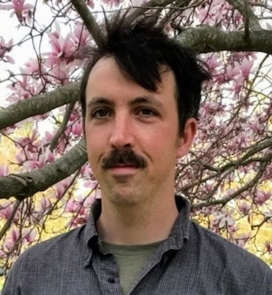 a person with short dark hair and a mustache stands in front of a blooming tree with pink flowers, wearing a gray shirt.