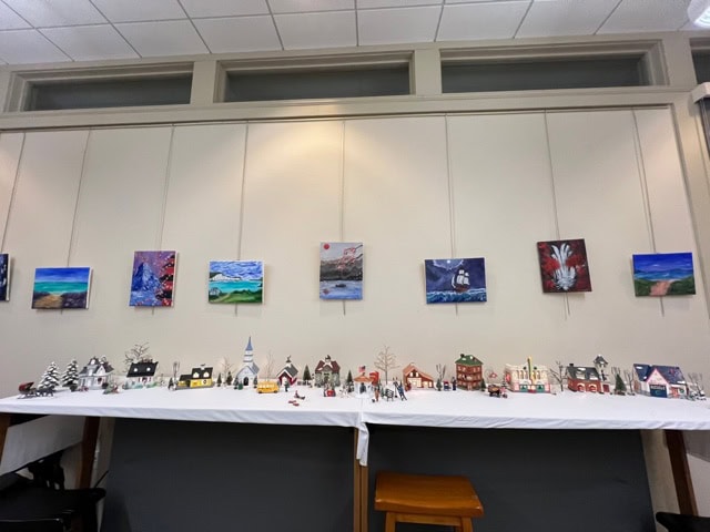 a table displays a miniature village scene with small houses and trees, set beneath a wall with framed landscape paintings.