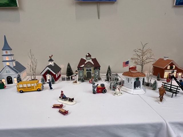 miniature village display features a school bus, small buildings, trees, figurines, a horse, and a gazebo with an american flag, all arranged on a white tablecloth.