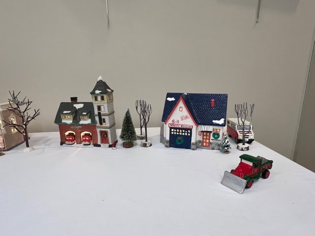miniature holiday village display with small model houses, leafless trees, a christmas tree, and a toy snowplow on a white tablecloth background.