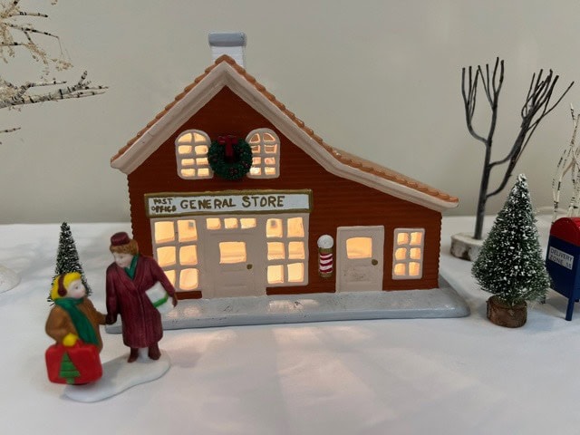 a miniature model of a general store with lit windows, two small figurines in winter clothes, artificial trees, and a mailbox in a snowy setting.