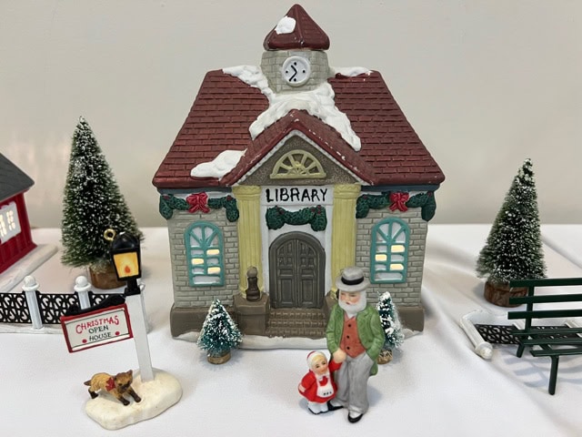 a miniature model of a library with snow, holiday decorations, two figurines, trees, a bench, and a "christmas open house" sign in front.