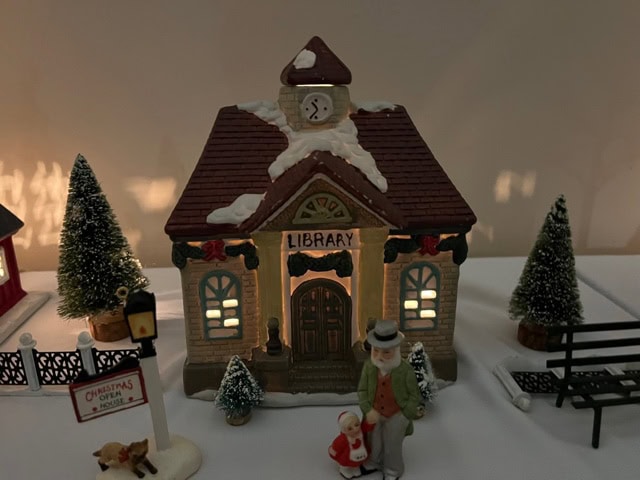 a miniature model of a snow covered library with lit windows, a clock above the door, and figurines of an elderly man and a child standing in front.