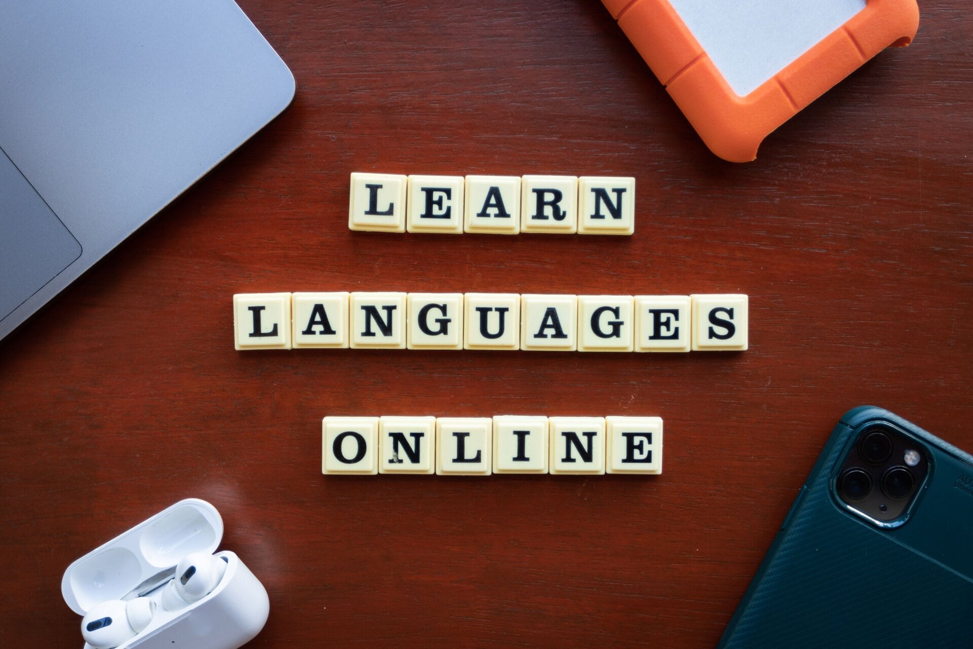 letter tiles spell “learn languages online” on a wooden table, surrounded by a laptop, earphones, an external drive, and a smartphone.
