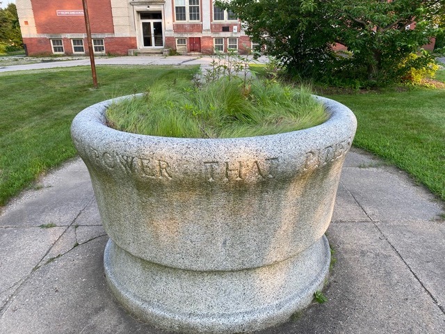 large stone planter with overgrown grass, engraved with partially visible text, sits on a concrete walkway in front of a brick building.