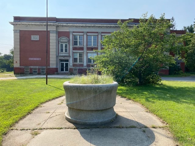 a round stone planter with overgrown grass stands on a concrete path in front of an old brick and stone school building.