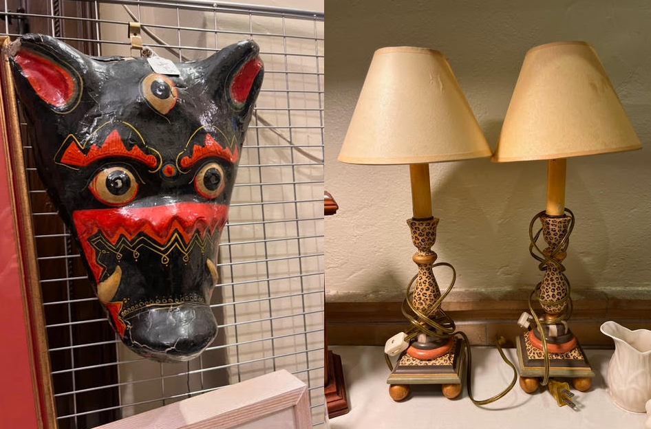 a painted animal mask with bold patterns hangs on a grid wall beside a pair of vintage table lamps with cream colored shades on a wooden surface.