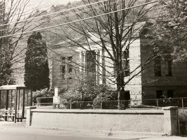 a stone building with columns and a fenced yard is partially obscured by trees; a bus stop shelter sits on the sidewalk in front.