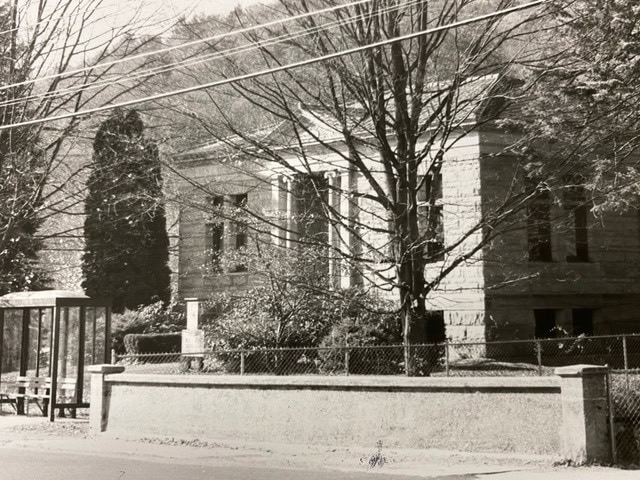 a stone building with columns and a fenced yard is partially obscured by trees; a bus stop shelter sits on the sidewalk in front.