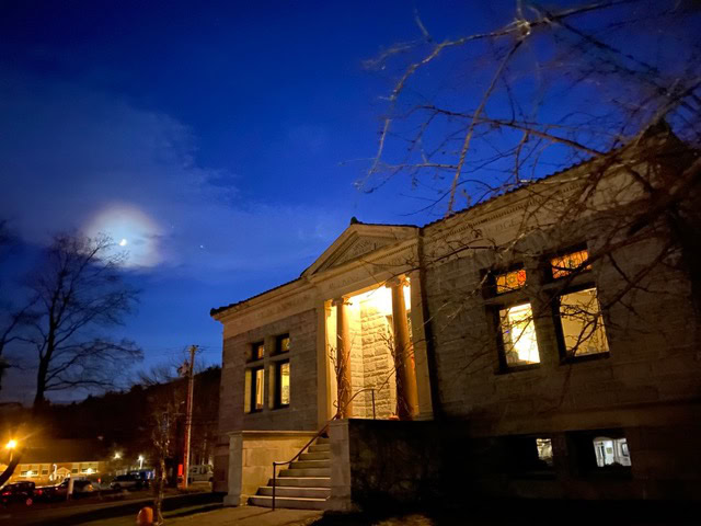 a stone building with lit windows and front steps is pictured at night under a dark blue sky with the moon partially visible behind clouds.