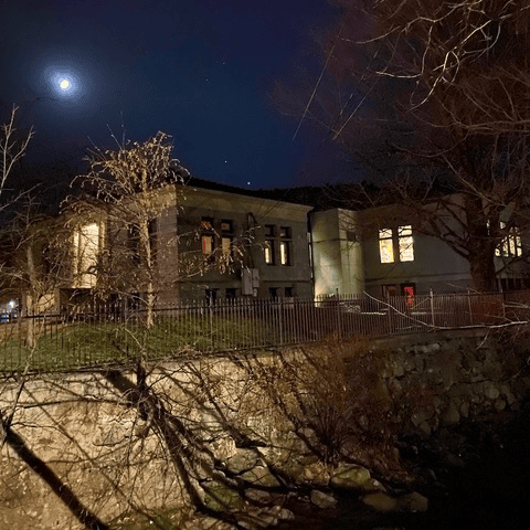 a building with illuminated windows stands behind a metal fence at night, with bare trees in the foreground and a crescent moon visible in the sky.