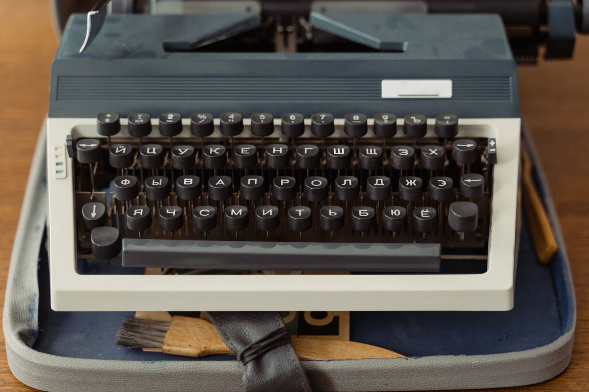 a vintage typewriter with black keys sits open on a table, with a brush and carrying case partially visible beneath it.