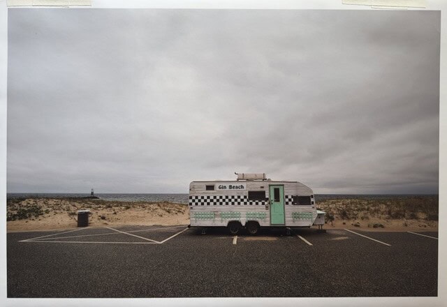 a food trailer labeled “gin beach” is parked in an empty parking lot near sand dunes under a cloudy sky.