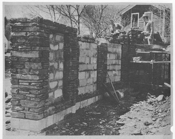 a worker stands on scaffolding while constructing a stone and brick wall outdoors, with a house and bare trees visible in the background.