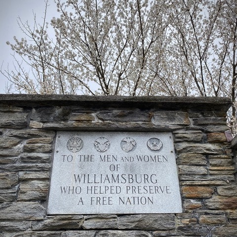 stone memorial plaque reads: "to the men and women of williamsburg who helped preserve a free nation," with military emblems above, set in a stone wall with blossoming trees behind.