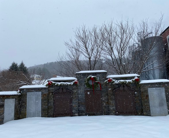 a stone memorial wall with plaques and wreaths is shown in a snowy outdoor setting, with bare trees and metal silos in the background.