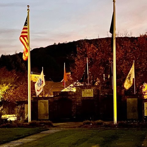 a stone memorial with multiple flagpoles and flags, including the american flag, stands in front of autumn trees at dusk.